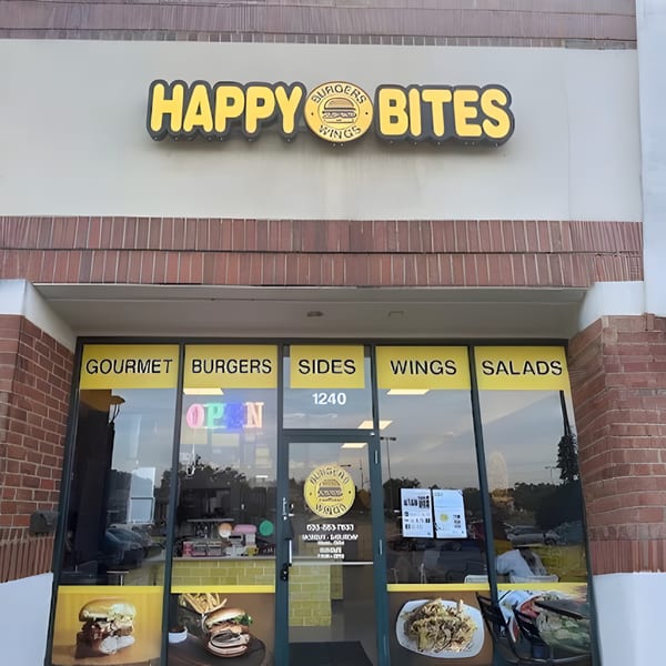 The storefront of Happy Bites restaurant, featuring a yellow sign and window decals advertising burgers, wings, and salads.